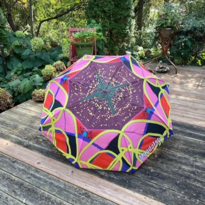 An umbrella on a wood deck, featuring a custom red, pink, yellow, and purple mandala design.
